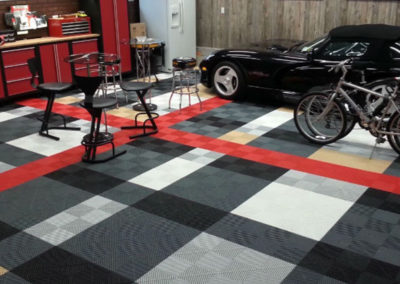 A garage with a decorative checkered floor, a black car, bicycles, red cabinets, and automotive-themed artwork on the walls.
